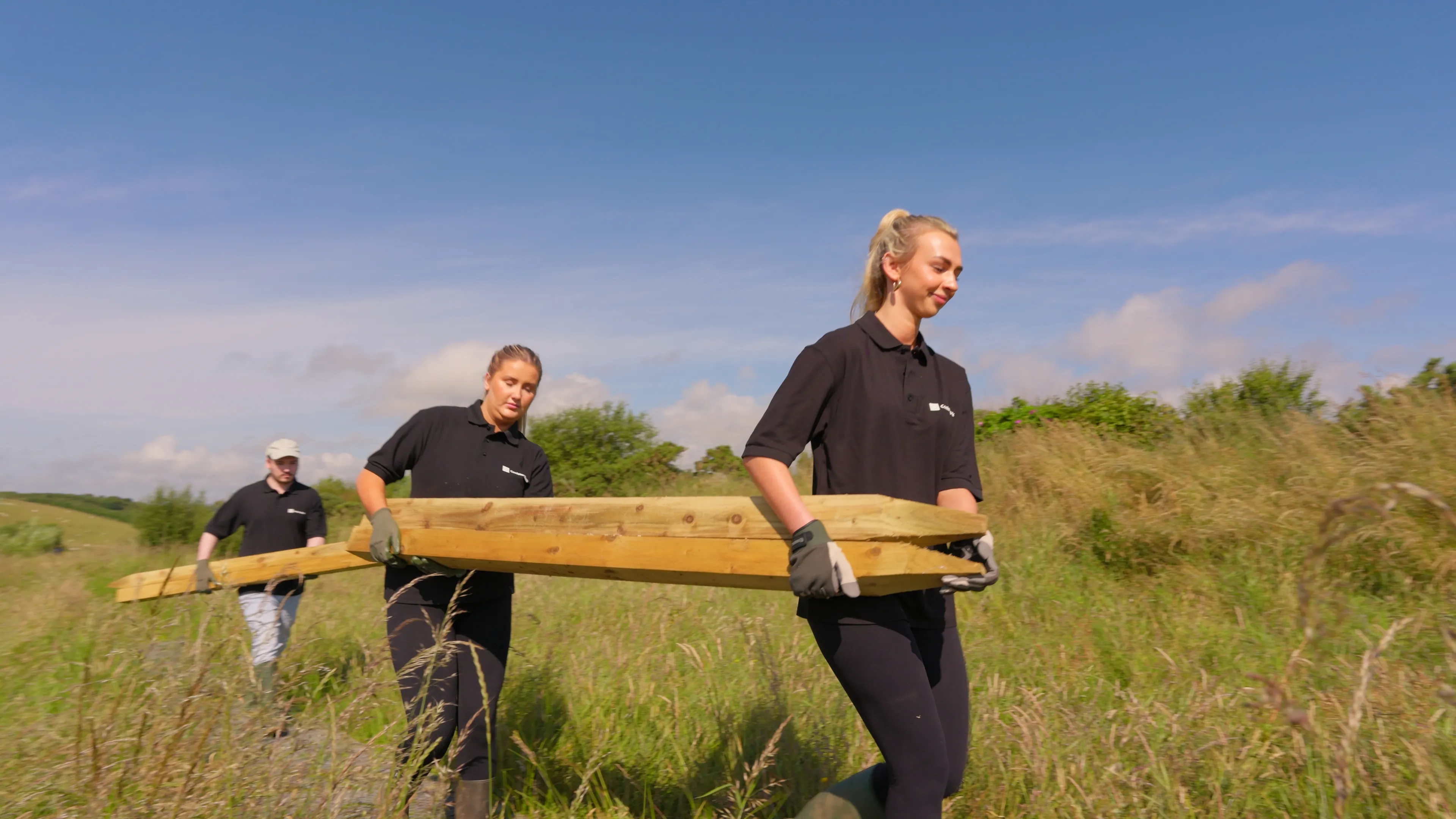 Conister Bank staff carrying timber across open ground during the bee safe garden volunteer week at The Children's Centre, Isle of Man, filmed by DotPerformance